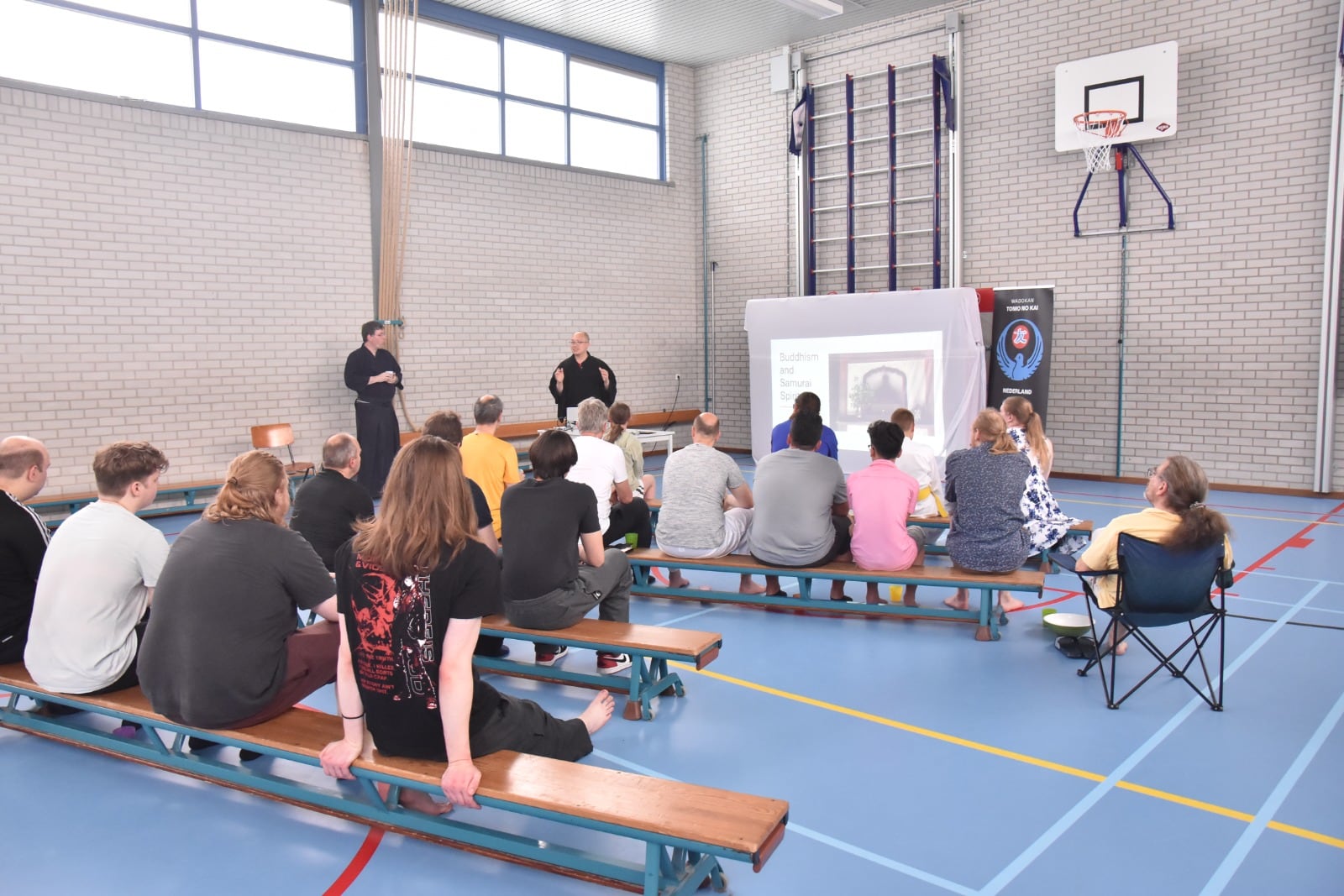 De leerlingen luisteren op de banken naar de presentatie in de dojo
